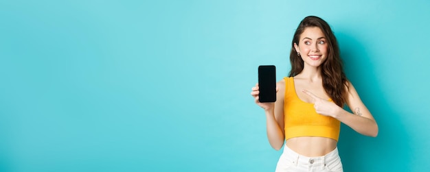 Kostenloses Foto porträt einer süßen brünetten frau, die mit dem finger auf den smartphone-bildschirm zeigt und nach links auf den kopierraum blickt, der im sommeroutfit vor blauem hintergrund steht