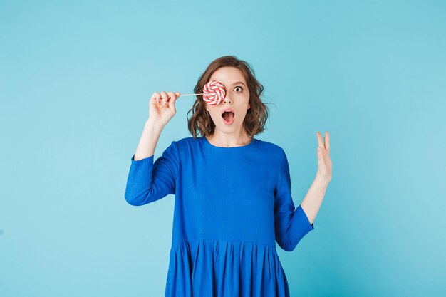 Porträt einer süßen Dame im Kleid, die steht und ihr Gesicht mit Lollipop-Süßigkeiten bedeckt, während sie verblüfft in die Kamera auf blauem Hintergrund blickt