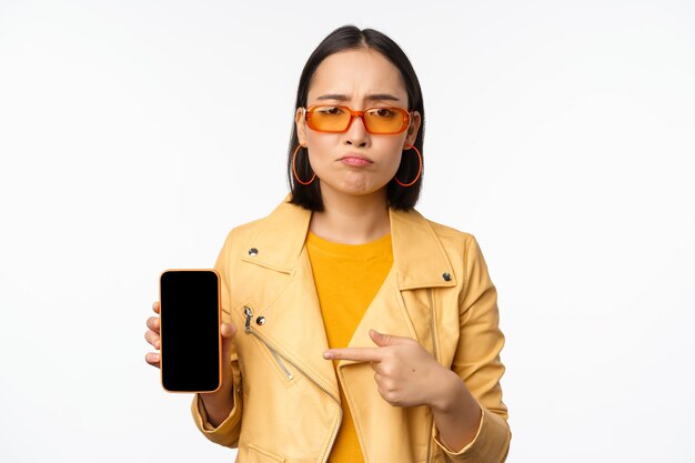 Porträt einer traurigen asiatischen Frau mit Sonnenbrille, die mit dem Finger auf die Schnittstelle der Handy-App zeigt und die Smartphone-Anwendung zeigt, die über weißem Hintergrund steht