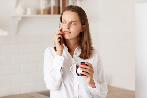 Porträt einer traurigen nachdenklichen Frau, die am Handy spricht, eine Tasse hält, mit verärgertem Ausdruck wegschaut, in der Küche posiert und einen lässigen Stil trägt.