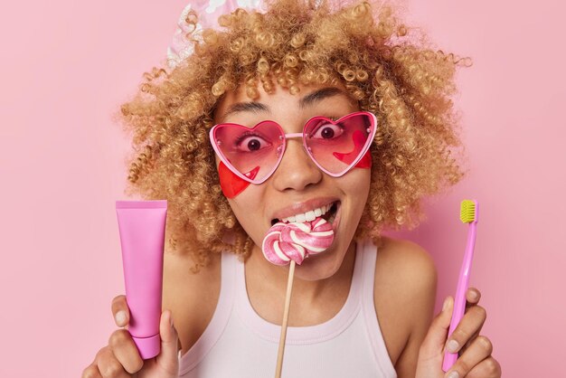 Porträt einer überraschten, fröhlichen Frau mit lockigem, buschigem Haar, hält Lutscher im Mund, hält Zahnpasta und Zahnbürste, trägt Schönheitsflecken unter den Augen auf, trägt rosafarbene Herz-Sonnenbrillen, Posen im Innenbereich