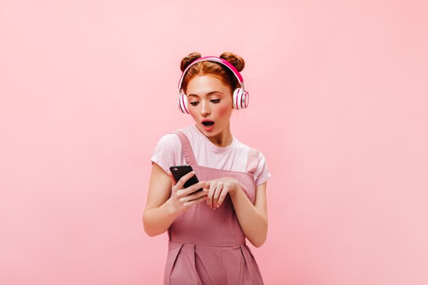 Porträt einer überraschten grünäugigen Frau im rosa Kleid. Frau, die Smartphone hält und Lieder in Kopfhörern hört.