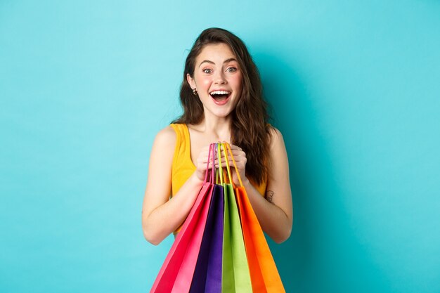 Porträt einer überraschten jungen Frau, die erstaunt in die Kamera schaut, Einkaufstüten hält, Rabatte im Laden sieht, auf blauem Hintergrund steht