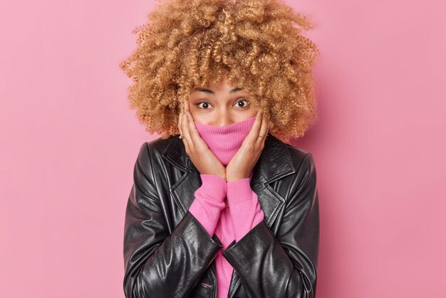 Porträt einer überraschten positiven jungen Frau mit lockigem Haar bedeckt den Mund mit Kragen in Lederjacke gekleidet direkt in die Kameraposen vor rosafarbenem Hintergrund. Menschen und Emotionen Konzept