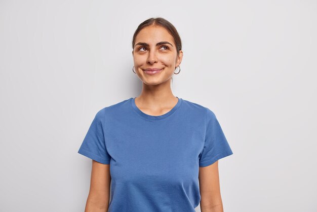 Porträt einer verträumten dunkelhaarigen Frau lächelt sanft konzentriert über etwas sehr Angenehmes, erinnert sich an schöne Erinnerungen, trägt ein lässiges blaues T-Shirt isoliert über weißer Studiowand