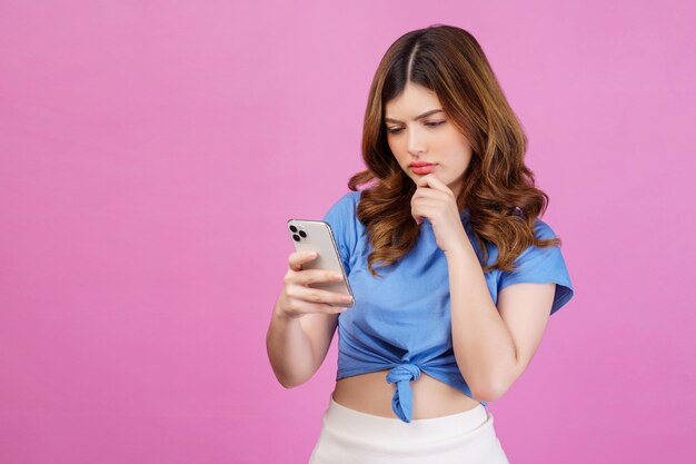 Porträt einer verwirrten jungen Frau, die ein lässiges T-Shirt mit einem Smartphone trägt, das über rosafarbenem Hintergrund isoliert ist