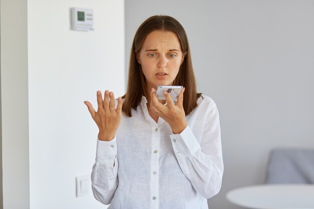 Porträt einer verwirrten verwirrten Frau mit weißem Hemd, die zu Hause mit Smartphone in den Händen posiert, den Arm hebt, versteht nicht, warum das Gerät nicht funktioniert.