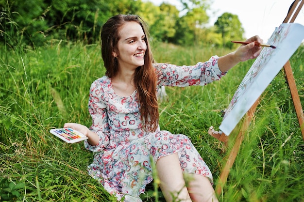 Porträt einer wunderschönen, glücklichen jungen Frau in schönem Kleid, die auf dem Gras sitzt und mit Wasserfarben auf Papier malt