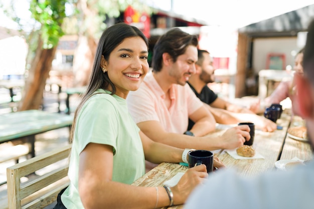 Porträt einer wunderschönen jungen Frau, die lächelt und Blickkontakt herstellt, während sie mit Menschen in einem Café im Freien sitzt. Brunch mit Freunden genießen