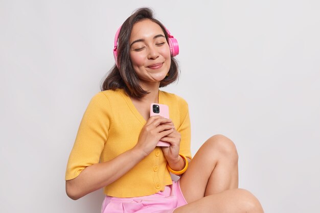 Porträt einer zufriedenen asiatischen Frau mit dunklem Haar genießt Lieblingsmusik perfekter Klang in drahtlosen Kopfhörern hält modernes Mobiltelefon hält die Augen geschlossen trägt Freizeitkleidung isoliert über weißer Wand