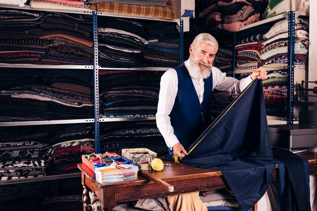 Porträt eines älteren männlichen Schneiders, der Maß des blauen Gewebes mit messendem Band auf Tabelle in seinem Shop nimmt