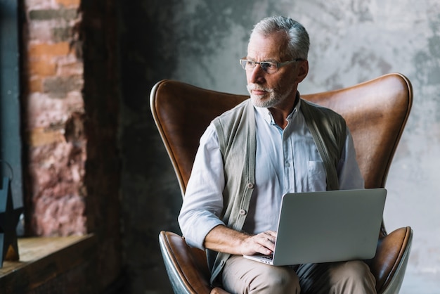 Porträt eines älteren Mannes, der auf Stuhl mit dem Laptop weg schaut sitzt