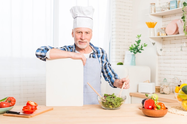 Porträt eines älteren Mannes, der das Halten des grünen Salats vorbereitet, überreichen das weiße Plakat in der Küche