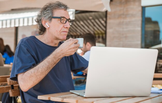 Porträt eines älteren Mannes, der die Kaffeetasse sitzt am Café im Freien mit Laptop hält