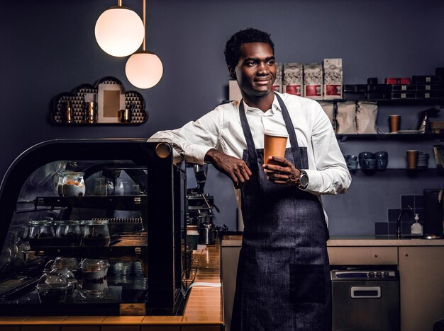 Porträt eines afrikanischen Baristas, der eine Tasse mit Kaffee hält, während er sich an eine Theke in einem Café lehnt