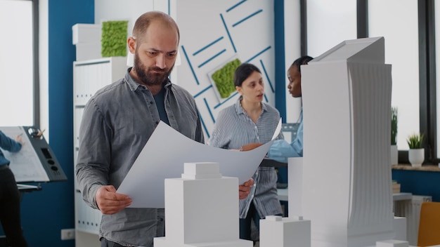 Porträt eines Architekten, der sich Blaupausenplan und Gebäudemodell auf dem Tisch ansieht, um den Bauplan und das Architekturprojekt zu entwerfen. Ingenieurplanung Stadtentwicklung mit Maquette