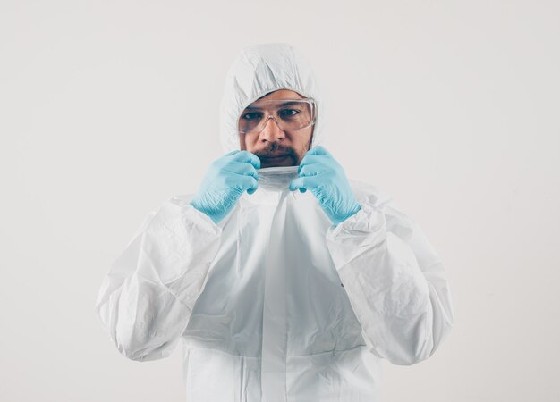 Porträt eines Arztes am hellen Hintergrund stehend und hält seinen Anzug in Maske, medizinischen Handschuhen und Schutzanzug.