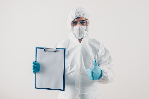 Porträt eines Arztes bei hellem Hintergrund, der medizinischen Bericht und Daumen in medizinischen Handschuhen und Schutzanzug hält