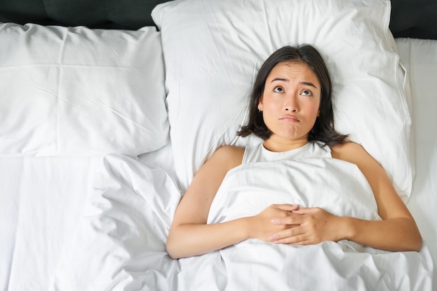 Porträt eines asiatischen Mädchens, das in ihrem Bett liegt und mit den Schultern zuckt, sieht verwirrt aus, bevor es schlafen geht