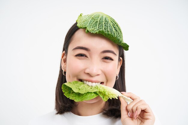 Porträt eines asiatischen Mädchens mit Blatt auf dem Kopf, das Kohl isst und isoliert auf weißem Hintergrund lächelt