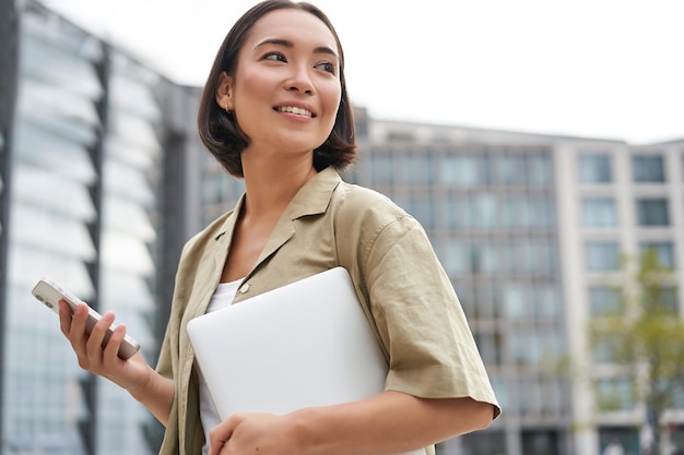 Porträt eines asiatischen Mädchens mit Laptop, das zur Arbeit oder zur Uni geht und ein Smartphone hält und lächelt, während es auf der Straße geht