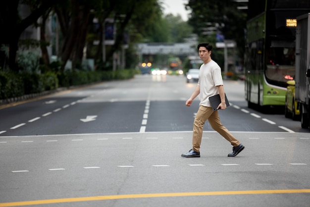 Porträt eines asiatischen Mannes, der die Straße in der Stadt überquert, während er einen Laptop hält