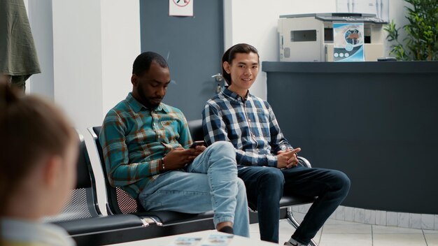 Porträt eines asiatischen Patienten, der mit Menschen in der Lobby des Wartebereichs sitzt und einen Arztbesuch beim Hausarzt an der Krankenhausrezeption hat. Wartezimmer in einer Gesundheitseinrichtung.