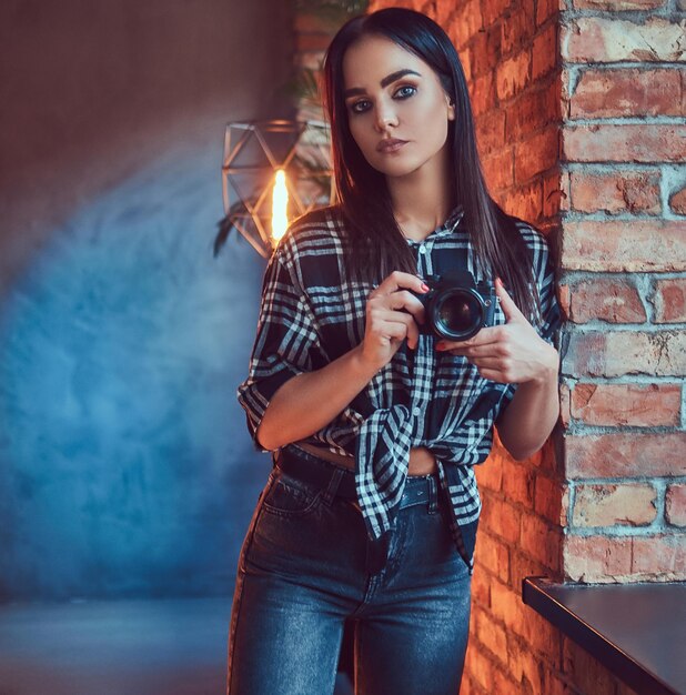 Porträt eines attraktiven brünetten Mädchens in Flanellhemd und Jeans mit Kamera an der Wand in einem Raum mit Loft-Interieur.