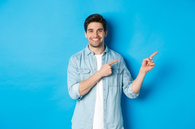 Porträt eines attraktiven erwachsenen Mannes, der lächelt, mit dem Finger direkt auf das Logo oder das Banner zeigt und Werbung vor blauem Hintergrund zeigt