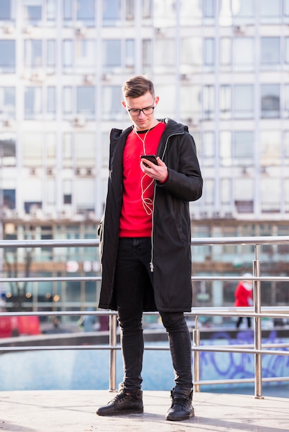 Porträt eines attraktiven jungen Mannes, der hörende Musik der langen Jacke auf Mobiltelefon trägt