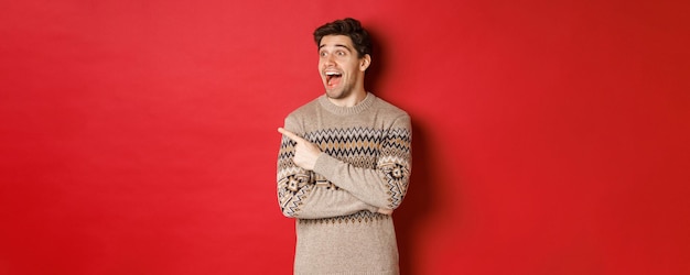 Porträt eines attraktiven kaukasischen Mannes, der die Winterferien des neuen Jahres feiert, mit dem Finger zeigt und mit erstauntem Ausdruck nach links schaut, der Weihnachtswerbung zeigt, die über rotem Hintergrund steht