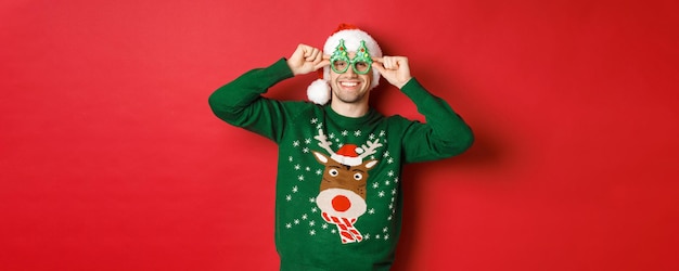 Porträt eines attraktiven lächelnden Mannes mit Weihnachtsmütze, Partybrille und Pullover, der Neujahrsferien feiert