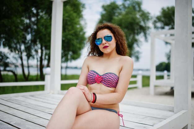 Porträt eines attraktiven Mädchens, das im Bikini mit Sonnenbrille posiert, während es auf einer weißen Holzterrasse steht