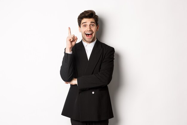 Porträt eines attraktiven Mannes im Anzug, der eine Idee hat, aufgeregt steht und einen Finger hebt, um Vorschlag, Denklösung zu erzählen, auf weißem Hintergrund stehend
