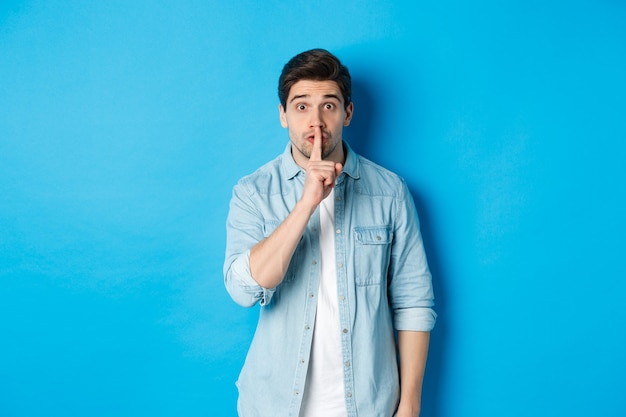 Porträt eines aufgeregten Mannes, der darum bittet, ruhig zu bleiben, ein Schweige-Tabu-Zeichen zeigt und nervös in die Kamera schaut, vor blauem Hintergrund stehend