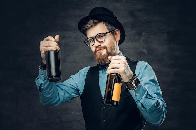 Kostenloses Foto porträt eines bärtigen hipster-männchens in einem blauen hemd, einer eleganten weste und einem zylinder hält zwei craft-bierflaschen mit einem gedanken an eine party.