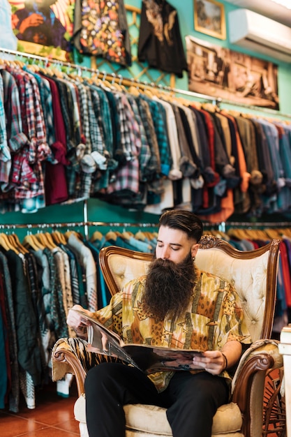 Kostenloses Foto porträt eines bärtigen mannes, der auf dem antiken sessel betrachtet zeitschrift im kleidungsshop sitzt