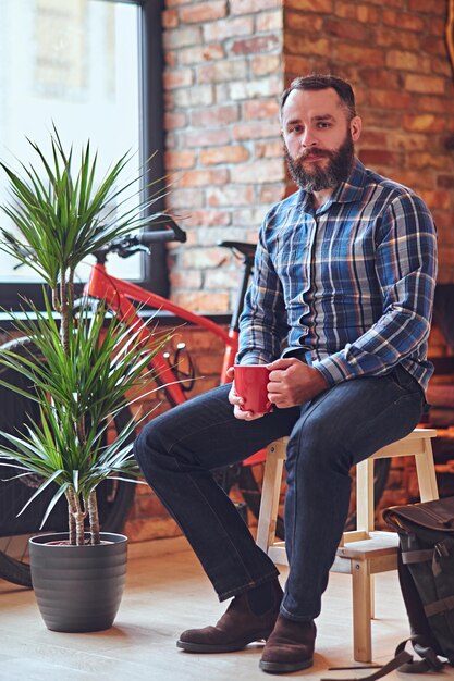 Porträt eines bärtigen Mannes in einem blauen Fleecehemd, trinkt heißen Kaffee in der Nähe des Fensters im Ruheraum mit Loft-Interieur.