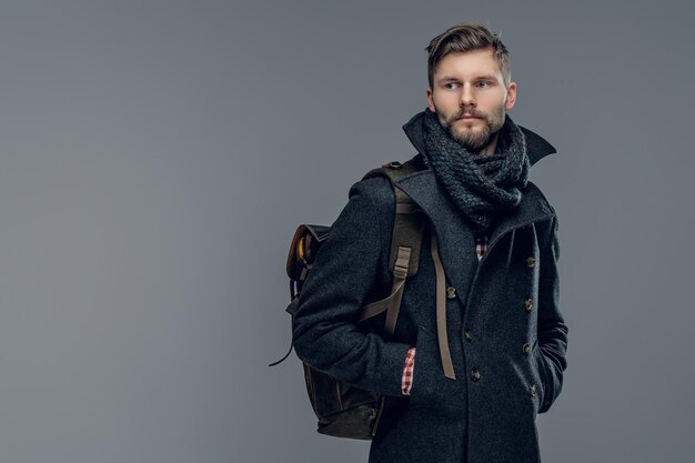 Porträt eines bärtigen Mannes in warmer Jacke und Schal hält einen Rucksack isoliert auf grauem Hintergrund.