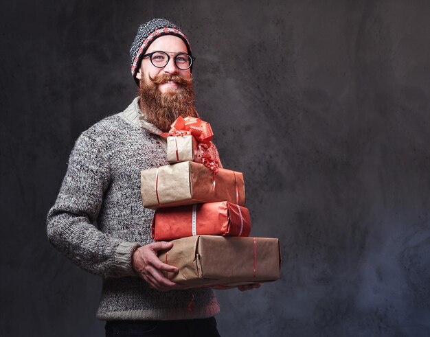 Porträt eines bärtigen Mannes mit Brille, gekleidet in einen warmen Wollpullover und einen Hut, hält Weihnachtsgeschenke auf grauem Hintergrund.