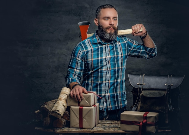 Porträt eines bärtigen Mannes mittleren Alters hält eine Axt und steht neben einem Tisch mit Weihnachtsgeschenkboxen und Brennholz auf grauem Hintergrund.