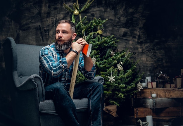 Porträt eines bärtigen Mannes mittleren Alters in kariertem Flanellhemd und Jeans sitzt auf einem Stuhl und hält eine Axt über weihnachtlich dekoriertem Hintergrund.