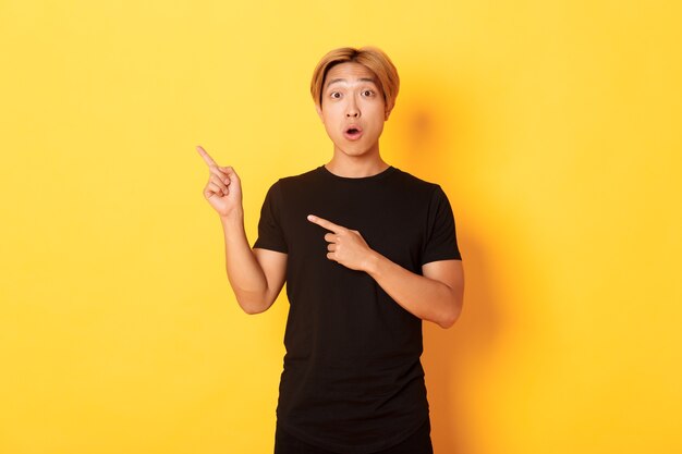 Porträt eines beeindruckten und aufgeregten gutaussehenden asiatischen Mannes im schwarzen T-Shirt, reagieren Sie auf Ihr Logo und zeigen Sie mit den Fingern in der oberen linken Ecke