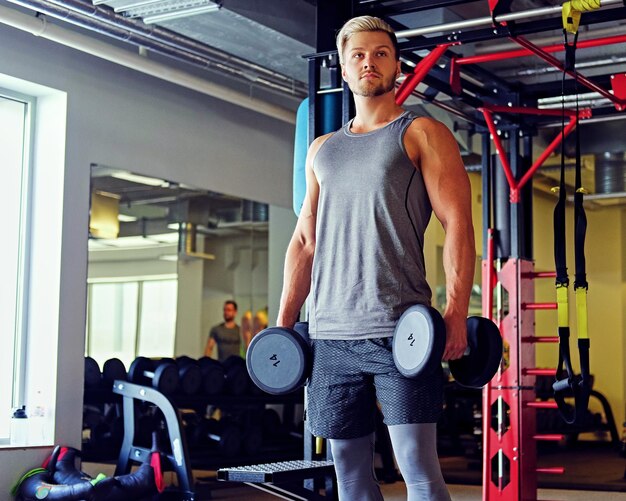 Porträt eines blonden sportlichen Mannes, der Kniebeugen mit Hanteln in einem Fitnessstudio macht.