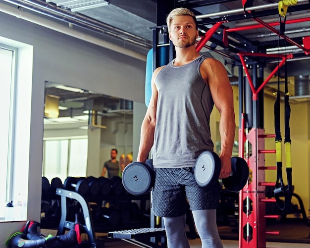 Porträt eines blonden sportlichen Mannes, der Kniebeugen mit Hanteln in einem Fitnessstudio macht.