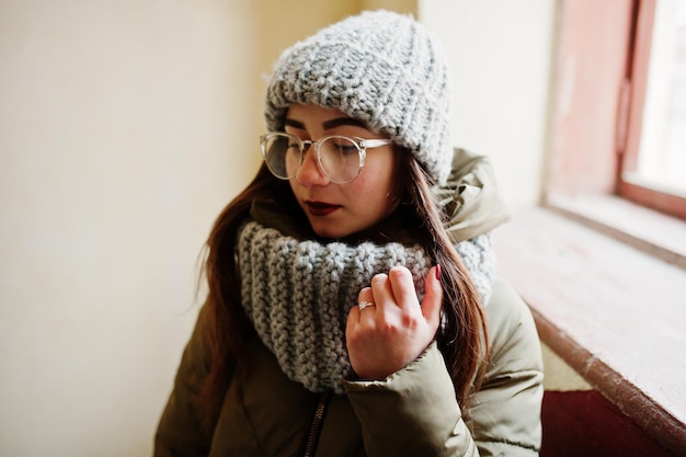 Porträt eines brünetten Mädchens mit grauem Schal und Hutbrille in der Nähe des Fensters