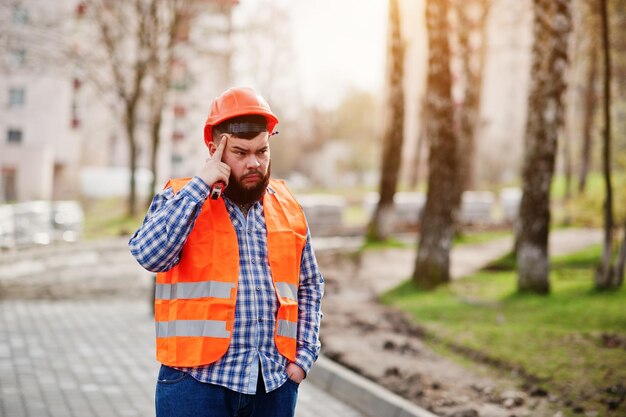 Porträt eines brutalen Bauarbeiters mit Bartarbeiter in einem orangefarbenen Sicherheitshelm gegen das Denken auf dem Bürgersteig