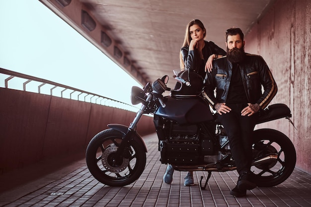 Porträt eines brutalen, modischen Bikers in einer schwarzen Lederjacke mit Sonnenbrille, der auf seinem maßgeschneiderten Retro-Motorrad sitzt, und seiner jungen brünetten Freundin, die sich an seine Schulter lehnt.