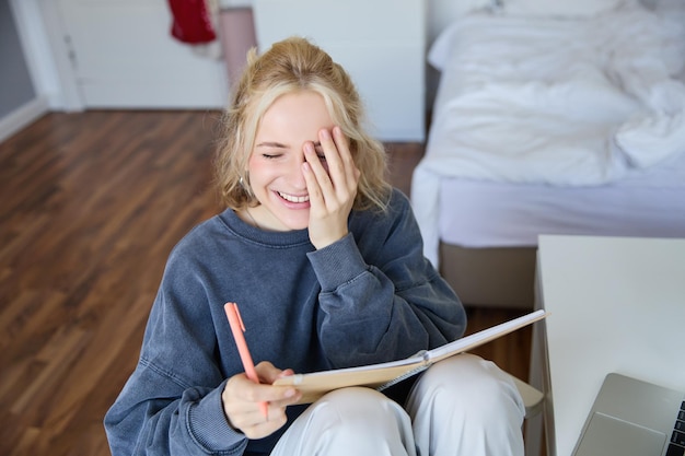 Kostenloses Foto porträt eines charismatischen blonden mädchens, eine lächelnde frau im schlafzimmer, die ein notizbuch und einen stift hält, in dem sie schreibt