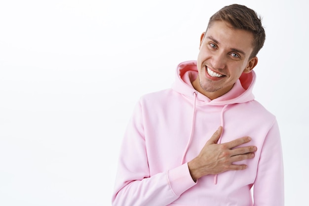 Kostenloses Foto porträt eines dankbaren, glücklichen blonden mannes in rosa hoodie, der sich für hilfe oder lob bedankt, das herz berührt und erfreut lächelt, dankbar oder froh ist, kamera schaut, rosa hoodie trägt, weiße wand